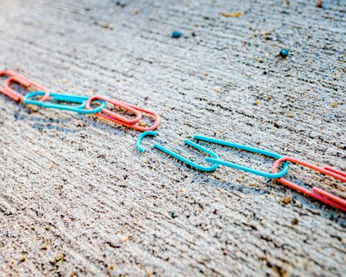 Red and Pink Paperclips are linked together on a concrete sidewalk; the center link of the "chain" is broken